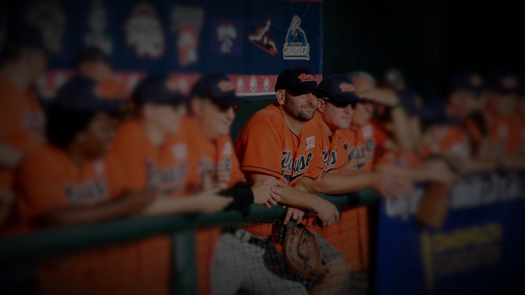 Softball players in orange jerseys lined up along the dugout rail